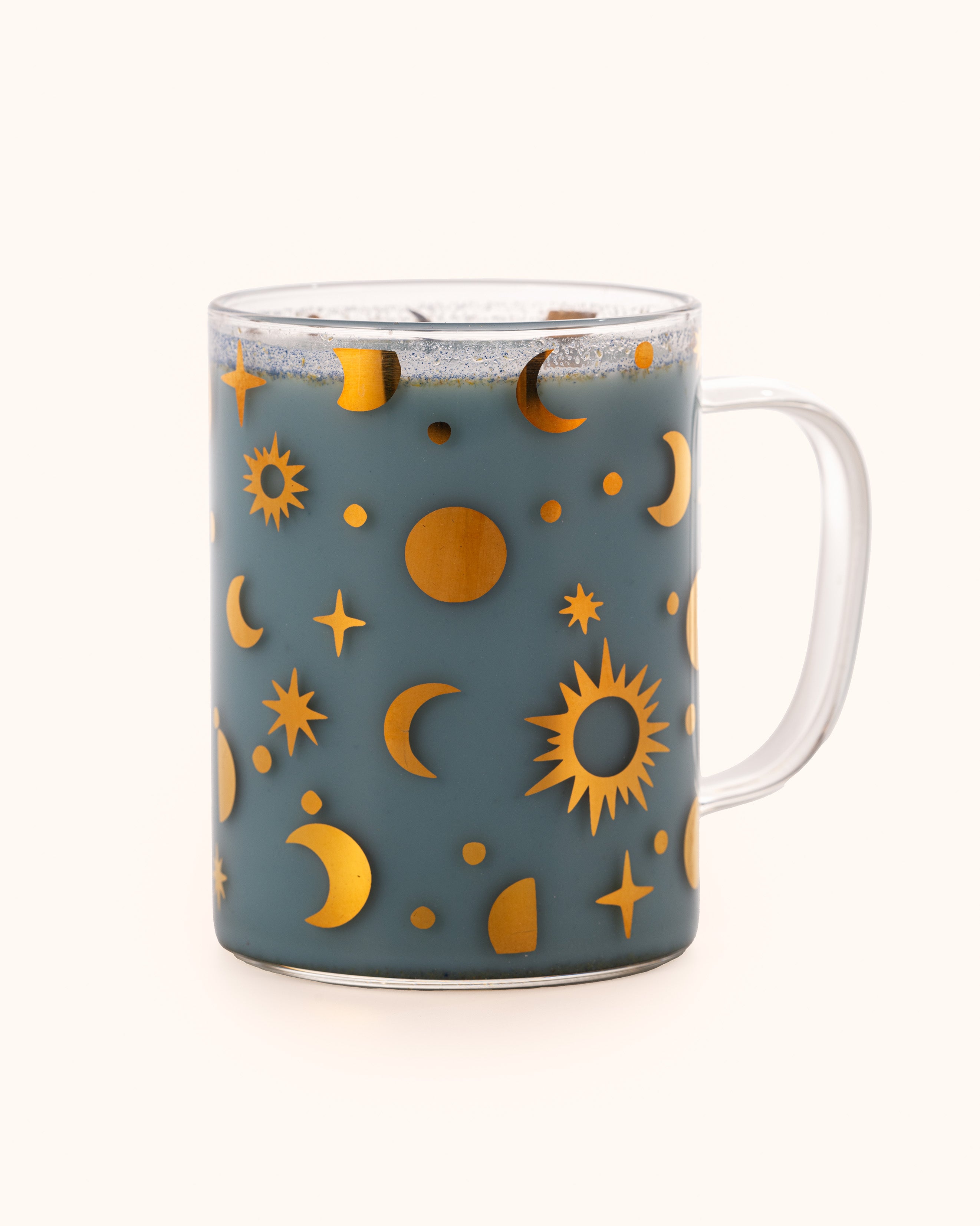 A clear glass mug featuring a pattern of shimmering golden moons, suns, and stars, filled with Mama Thyme's Blue Moon Dream Milk; a beautiful baby blue beverage.