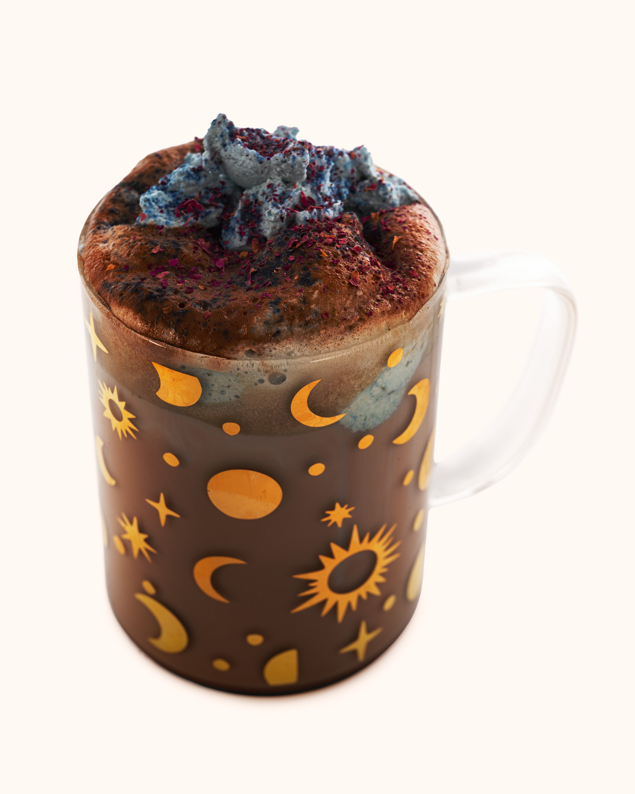 A clear glass mug featuring a pattern of shimmering golden moons, suns, and stars, filled with hot cocoa.