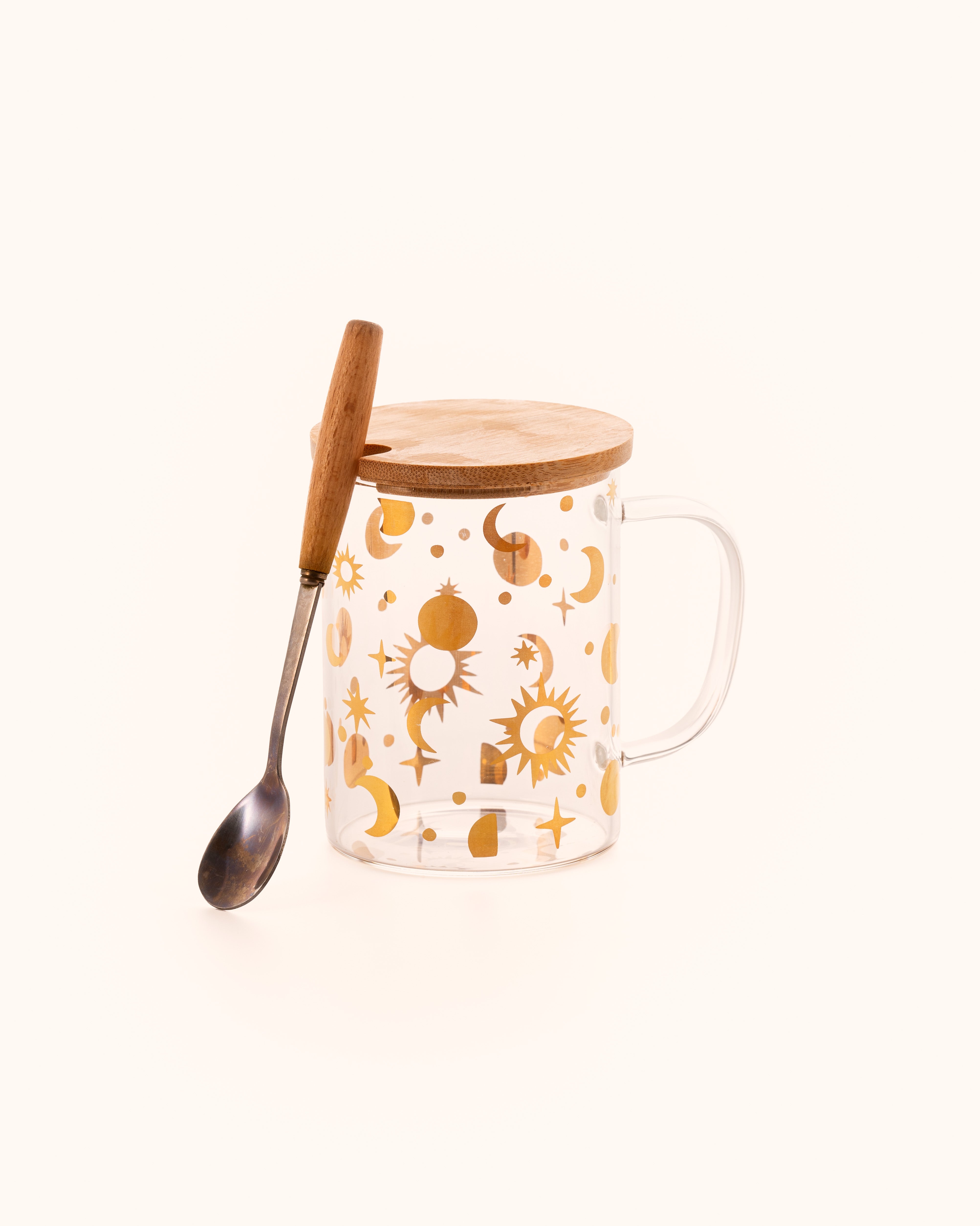 A clear glass mug featuring a pattern of shimmering golden moons, suns, and stars, with a wooden lid, and a metal stirring spoon that has a wooden handle.