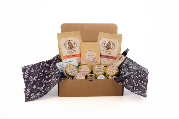A beautifully curated gift box open to reveal various packaged food and care products, accompanied by natural wood shavings and deep purple tissue paper featuring a floral pattern.